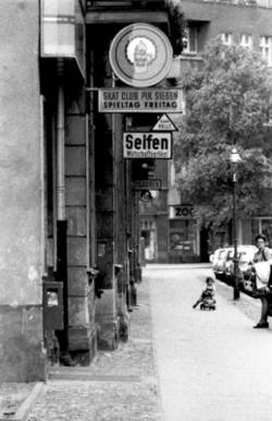 o.T., Straße mit diversen Werbeschildern: Skat Club PIK Sieben, Seifen, Zigarren; auf dem Bürgersteig Mutter mit Kind