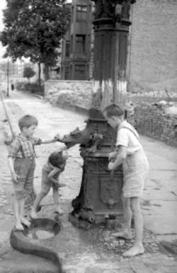 Kinder trinken aus einer Wasserpumpe am Rand der Straße.