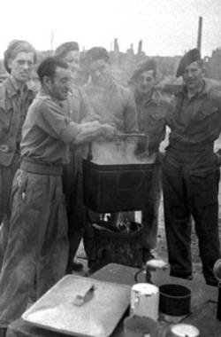 Mehrere Männer in Uniformen kochen etwas über einer Feuertonne.