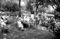 Kinder an Stühlen und Tischen in einem Wald mit einer Frau, die sie beaufsichtigt. Möglicherweise eine Waldschule