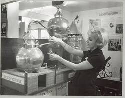 o.T., Am Stand einer Industrieaustellung bedient eine Frau einen Destillationsapparat