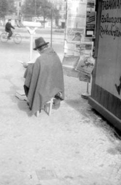 Ein sitzender Mann ließt eine Zeitung neben einen Zeitungsstand.