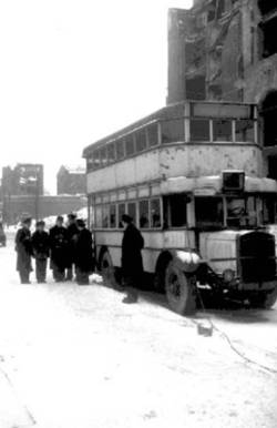 Straßenszene, Doppelstockbus hält auf der Straße, mehrere Busfahrer stehen daneben, im Hintergrund Ruinen.