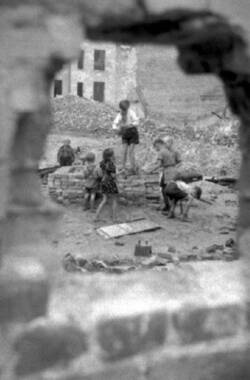 Blick durch ein Mauerloch auf zwischen Ruinen spielende Kinder