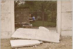 o.T., Blick durch eine herausgebrochene Hinterlandmaueröffnung auf eine in einem Garten sitzende Frau
